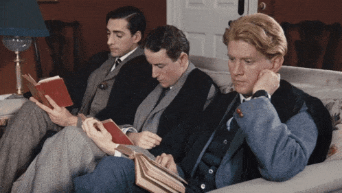 three men are sitting on a couch reading books and one is reading a red book