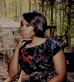 a woman in a floral dress is sitting in a chair with her hand on her chin in front of a sign that says ders