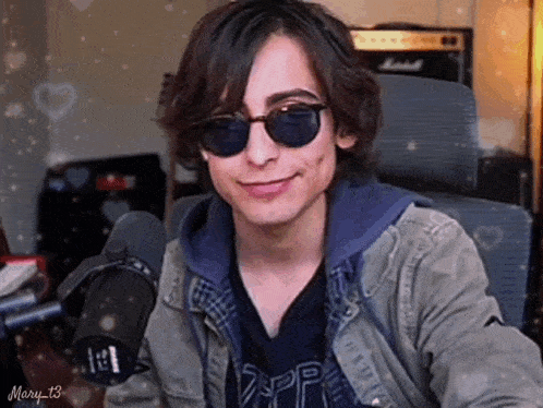 a young man wearing sunglasses and a jacket is sitting in front of a microphone and smiling .