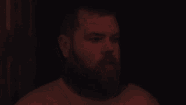 a man with a beard is covering his mouth with his hand while sitting in a dark room .