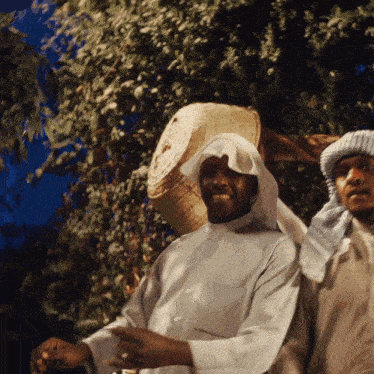 a man wearing a white shirt and a head scarf stands next to another man