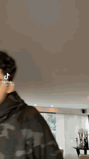 a man in a camouflage jacket is standing in a living room with a tik tok video .