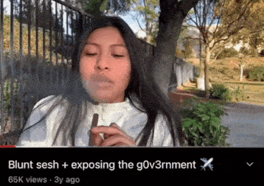 a woman is smoking a cigar in front of a fence and a tree .