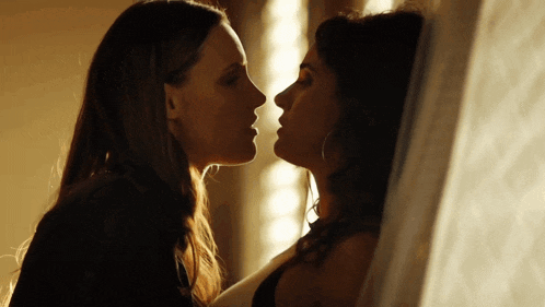 two women are kissing each other in front of a window in a dark room .