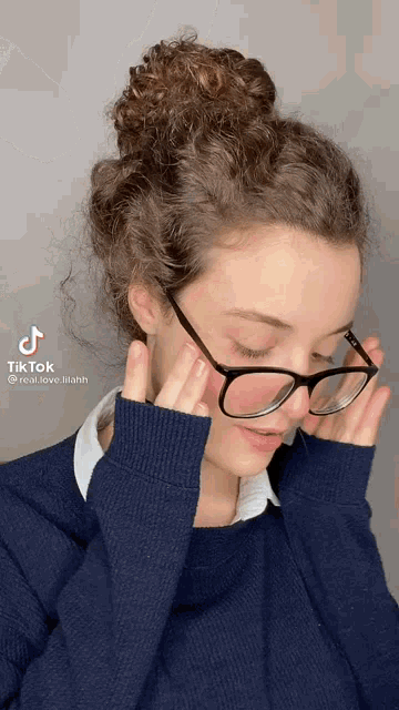 a young woman wearing glasses and a blue sweater is covering her face with her hands .
