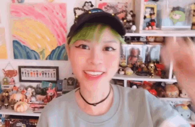 a woman with green hair wearing a hat and a choker is smiling in front of a shelf filled with toys .