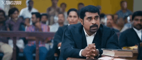 a man in a suit is sitting at a table with his hands folded in front of a group of people .