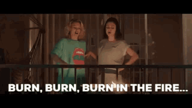 two women are standing on a balcony looking at a candle and saying `` burn , burn , burn in the fire . ''