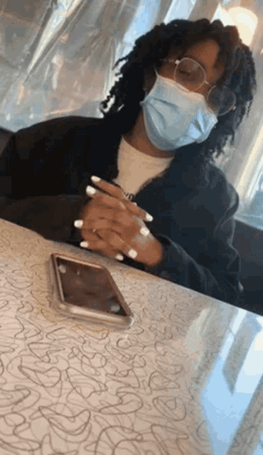 a woman wearing a face mask and glasses sits at a table with a cell phone