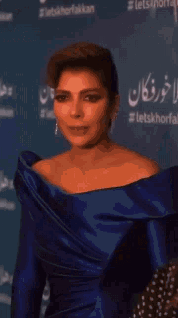 a woman in a blue off the shoulder dress stands in front of a sign that says letskhorfa