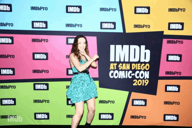 a woman stands in front of a colorful backdrop that says imdb at san diego comic con 2019