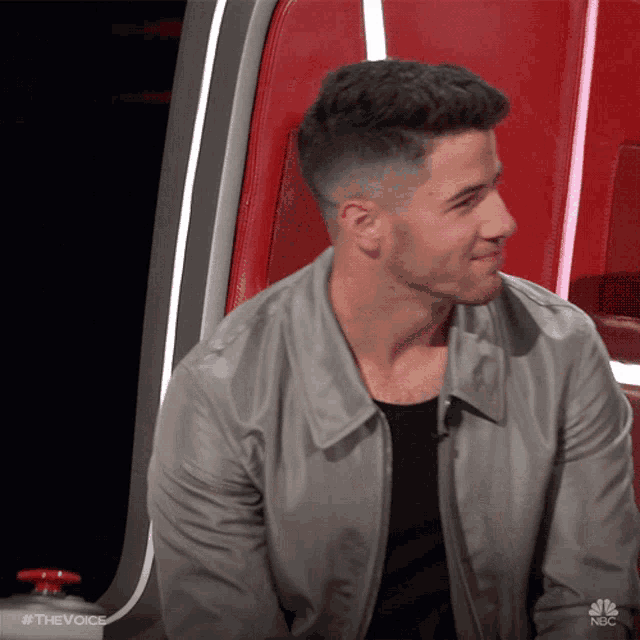 a man in a gray jacket is sitting in a red chair on the voice show