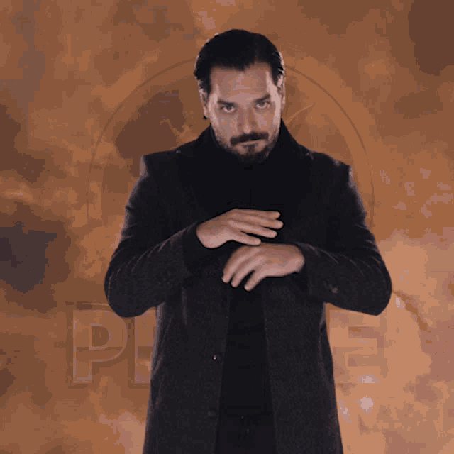 a man in a black coat stands with his hands on his chest in front of a cloudy background with the letter p on it