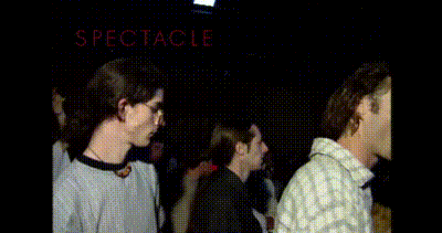 a man in a gray shirt is standing in a crowd with the word spectacle on the bottom right