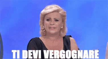 a woman in a black dress is sitting in front of a blue background with the words ti devi vergognare in white letters .