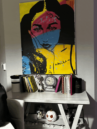 a colorful painting of a woman 's face hangs above a desk with books and a clock