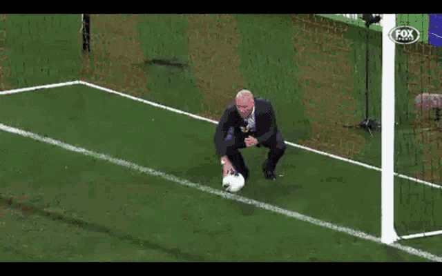 a man in a suit is kneeling on a soccer field with a soccer ball in his hand .