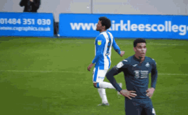 two soccer players on a field in front of a blue banner that says www.kirkleescollegiate