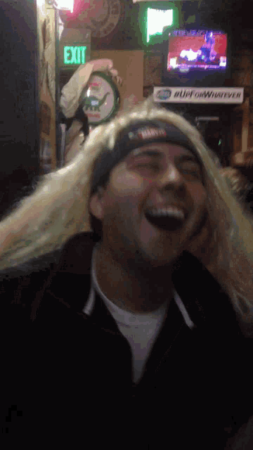 a man with long blonde hair is laughing in front of a green exit sign