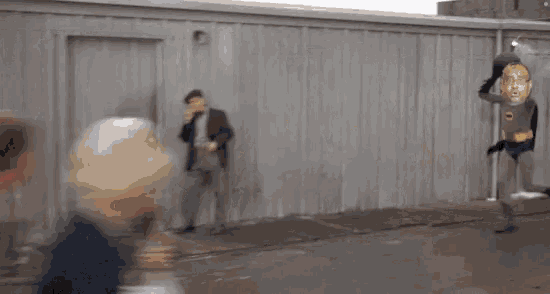 a man in a suit is standing in front of a building while a man in a batman costume is flying through the air