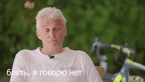 a man wearing a white t-shirt that says r.a. zlatka is sitting in front of a bicycle