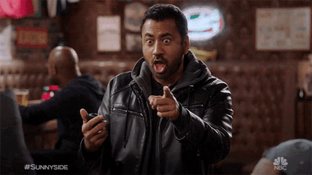 a man in a leather jacket is pointing at something while holding a cell phone with the nbc logo on the bottom right