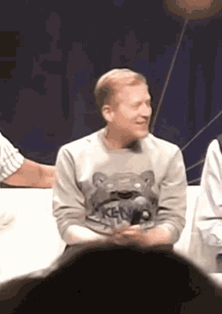 a man wearing a sweater with a bear on it is sitting on a couch and smiling .
