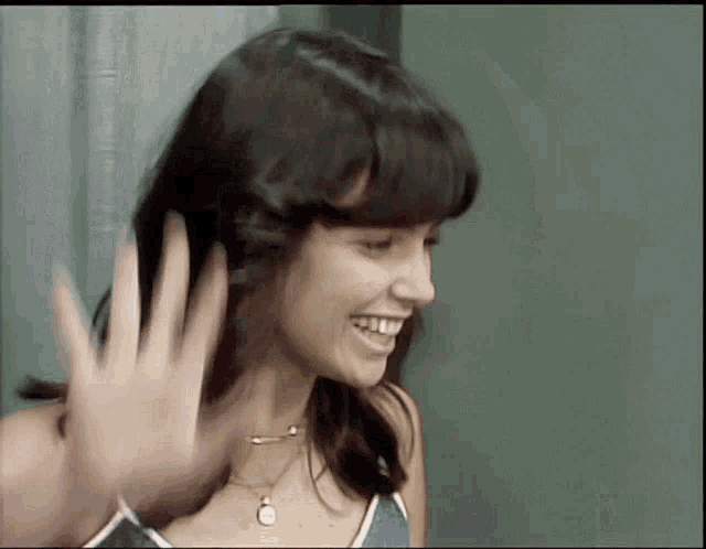 a woman is smiling and waving her hand in front of a green wall .