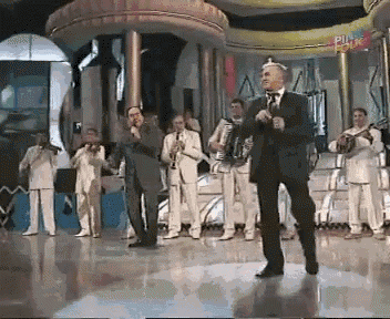 a man in a suit is singing into a microphone while a band plays instruments in front of him