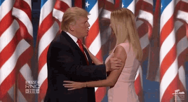 a man in a suit and tie is hugging a woman in a pink dress in front of american flags .