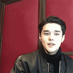 a young man is sitting in front of a red wall wearing a black jacket and turtleneck .