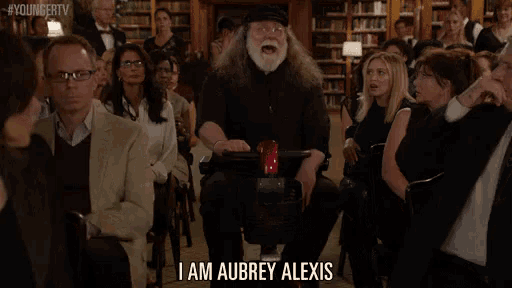 a man with long hair and a beard is standing in front of a crowd and says i am aubrey alexis