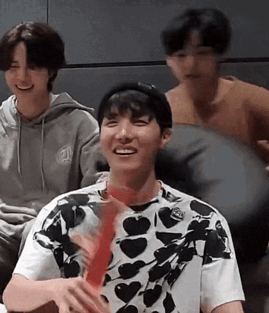 a group of young men are sitting on a couch and laughing . one of the men is wearing a heart print shirt .