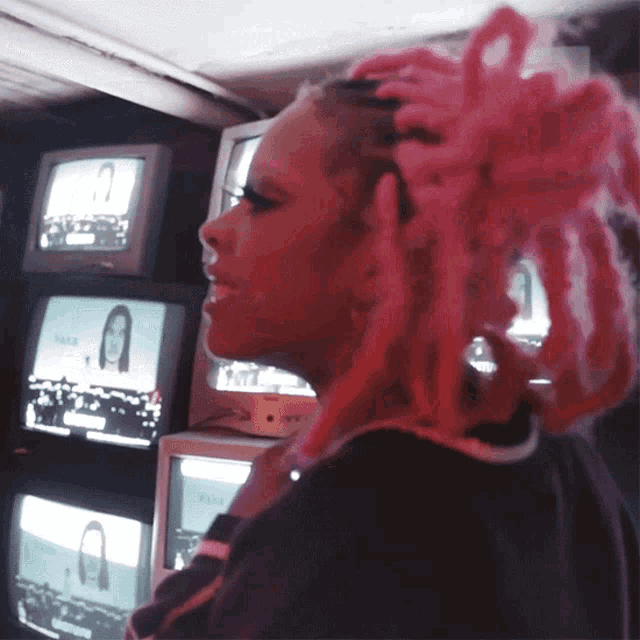 a woman with pink dreadlocks stands in front of a row of televisions including one that has a picture of a man on it