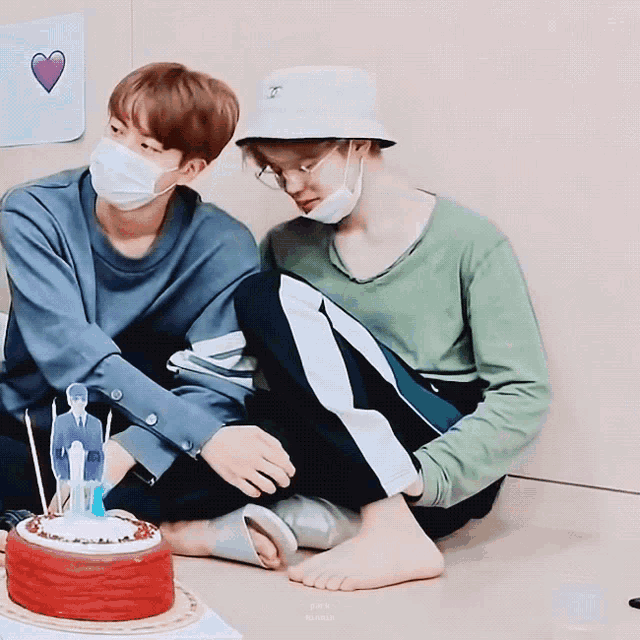 two men wearing face masks are sitting next to a red cake