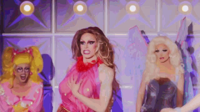 three drag queens are standing next to each other on a stage in front of a wall .