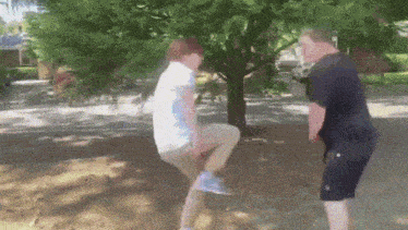 two men are squatting down under a tree in a park
