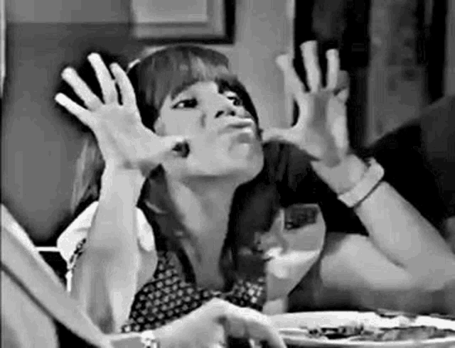 a woman is making a funny face with her hands while sitting at a table with a plate of food .