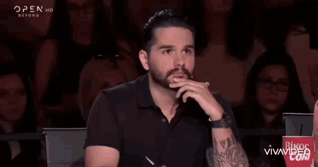 a man with a beard is sitting in front of a crowd in a theatre .