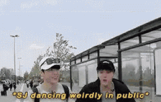 two men are standing next to each other in front of a bus stop and one of them is dancing weirdly in public .