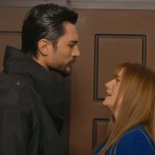 a man in a black jacket and a woman in a blue sweater look at each other