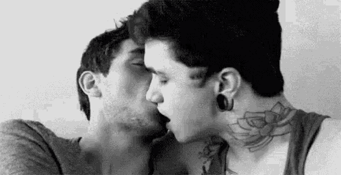 two men are kissing in a black and white photo . one of the men has a tattoo on his neck .