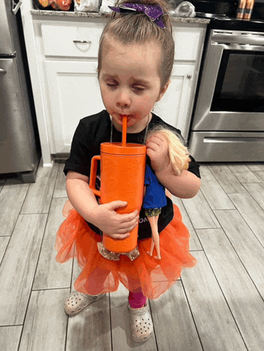 a little girl is drinking from an orange cup while holding a doll