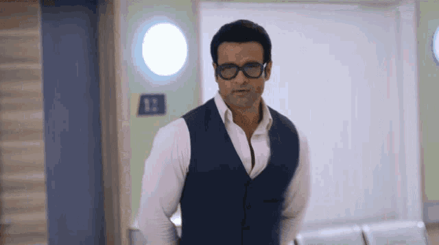 a man wearing glasses and a vest stands in a hallway with a sign that says 12
