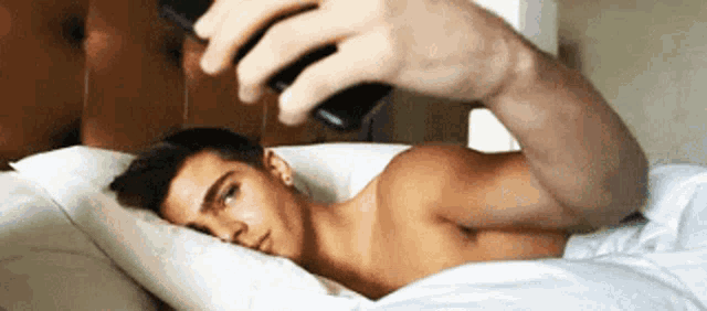 a shirtless man is laying in bed taking a selfie with his cell phone .