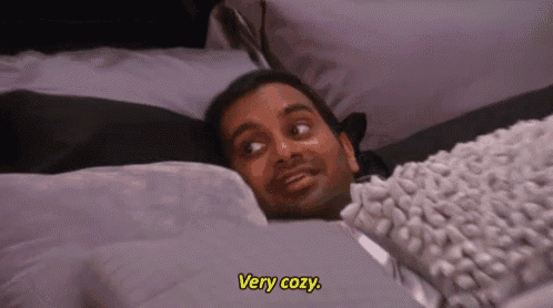 a man is laying on a bed with a blanket and a pillow and says `` very cozy '' .