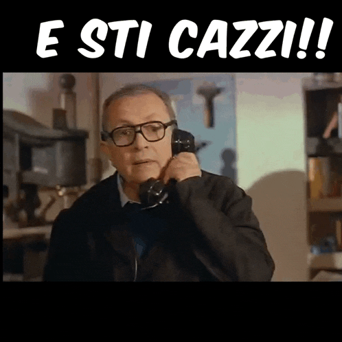 a man wearing glasses is talking on a phone with the words " e sti cazzi " behind him