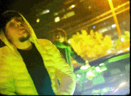 a man in a yellow jacket with a hood stands in front of a green car