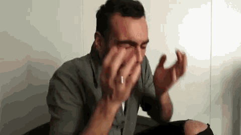 a man with a ring on his finger is sitting in a chair covering his eyes with his hands .