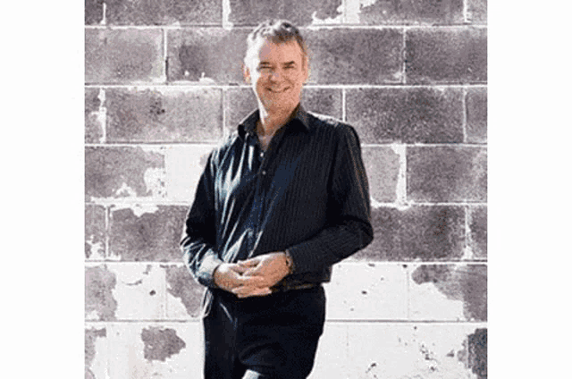 a man in a black shirt is standing in front of a brick wall and smiling .